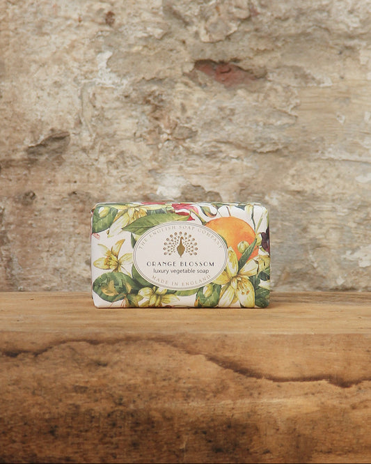Eine dekorative Tafel von ESC | 'Orange Blossom' pflanzliche Handseife mit fruchtigem Orangenblütenduft von The English Soap Company, verziert mit Blumen- und Fruchtmotiven, liegt auf rustikalem Holz vor einer Steinwand.