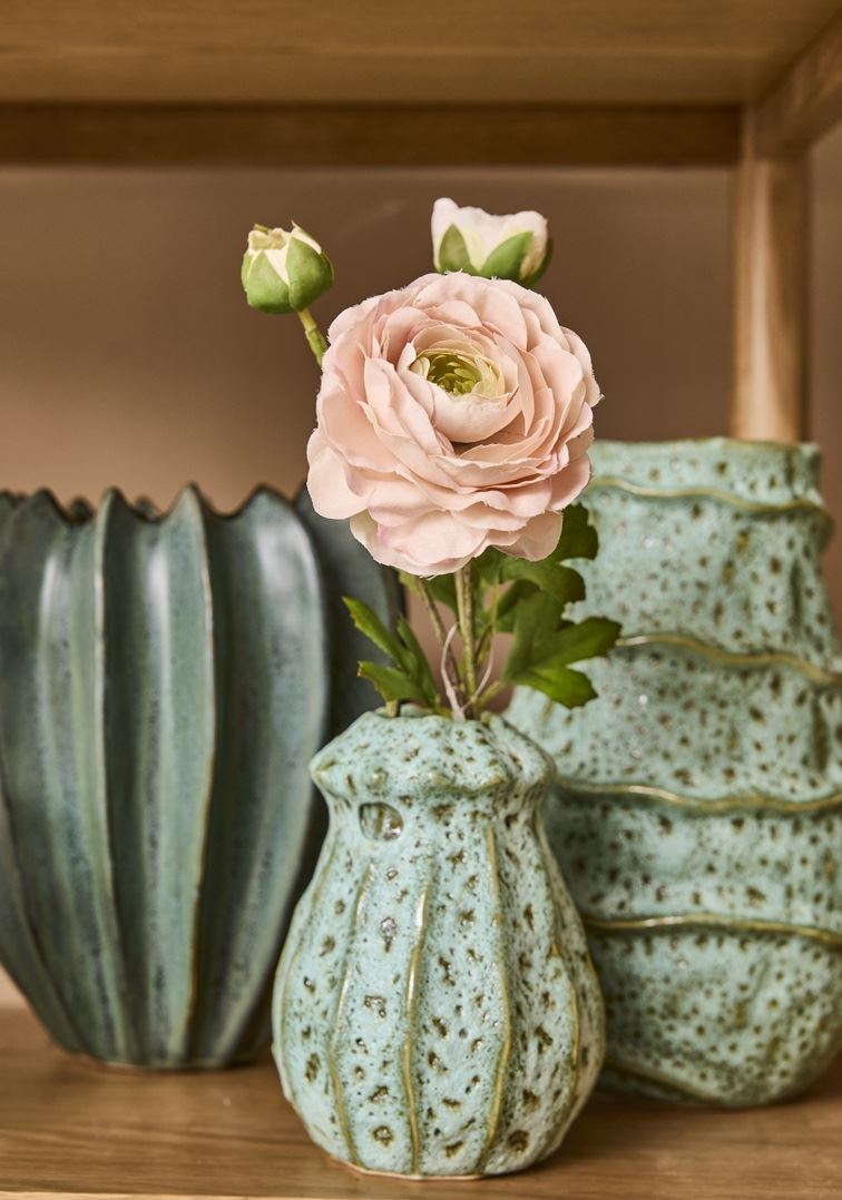 Eine hellrosa künstliche Ranunkel mit grünen Blättern ist in der 2 Have Vase 'Lavagrün' 15 x 11 cm arrangiert, die neben zwei größeren blau-grünen Keramikvasen auf einem Holzregal steht.