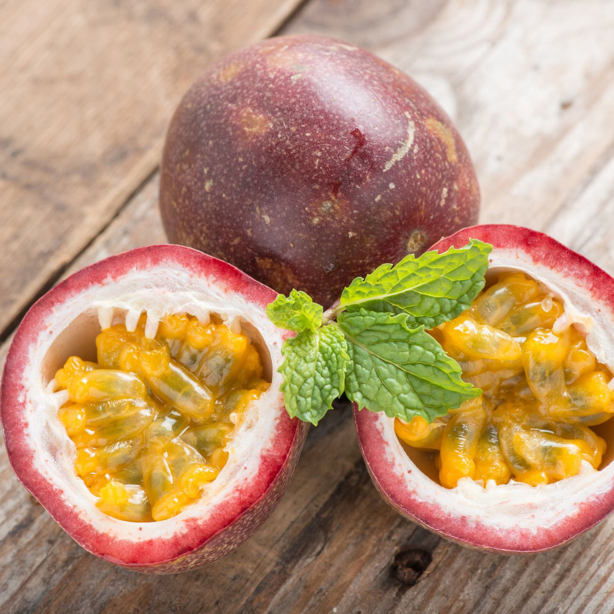 Eine ganze Passionsfrucht und eine halbierte Passionsfrucht, deren leuchtend gelbe Kerne und Fruchtfleisch neben einem Zweig frischer Minze auf einer rustikalen Holzunterlage glänzen – eine verlockende Präsentation, die an die lebhaften Aromen des „Wilkin & Sons Tiptree Passion Fruit Curd“ (312 g) erinnert.