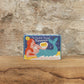 Eine kleine Schachtel mit The Printed Peanut | Rose Geranie Seife von The Printed Peanut steht auf einer hölzernen Oberfläche, hinter der sich eine strukturierte Steinwand befindet. Die Verpackung zeigt ein skurriles Design vor einem blauen Hintergrund.