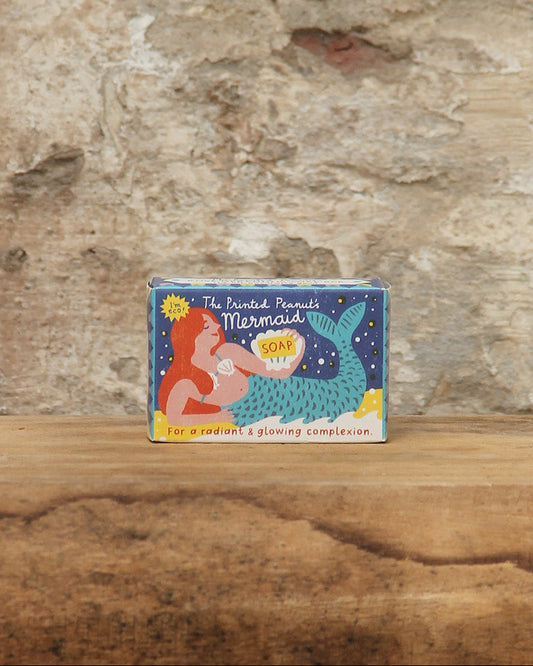 Eine kleine Schachtel mit The Printed Peanut | Rose Geranie Seife von The Printed Peanut steht auf einer hölzernen Oberfläche, hinter der sich eine strukturierte Steinwand befindet. Die Verpackung zeigt ein skurriles Design vor einem blauen Hintergrund.