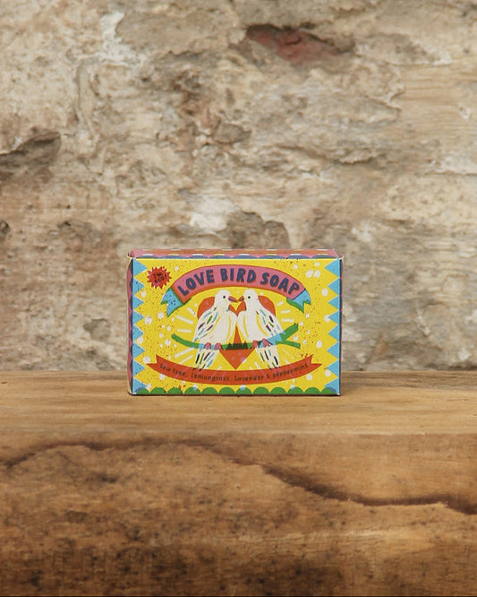 Eine farbenfrohe Schachtel von The Printed Peanut | Teebaumöl, Zitronengras, Lavendel und Pfefferminze Seife von The Printed Peanut mit zwei Vögeln, die sich gegenüberstehen, steht auf Holz vor einer strukturierten Steinwand.