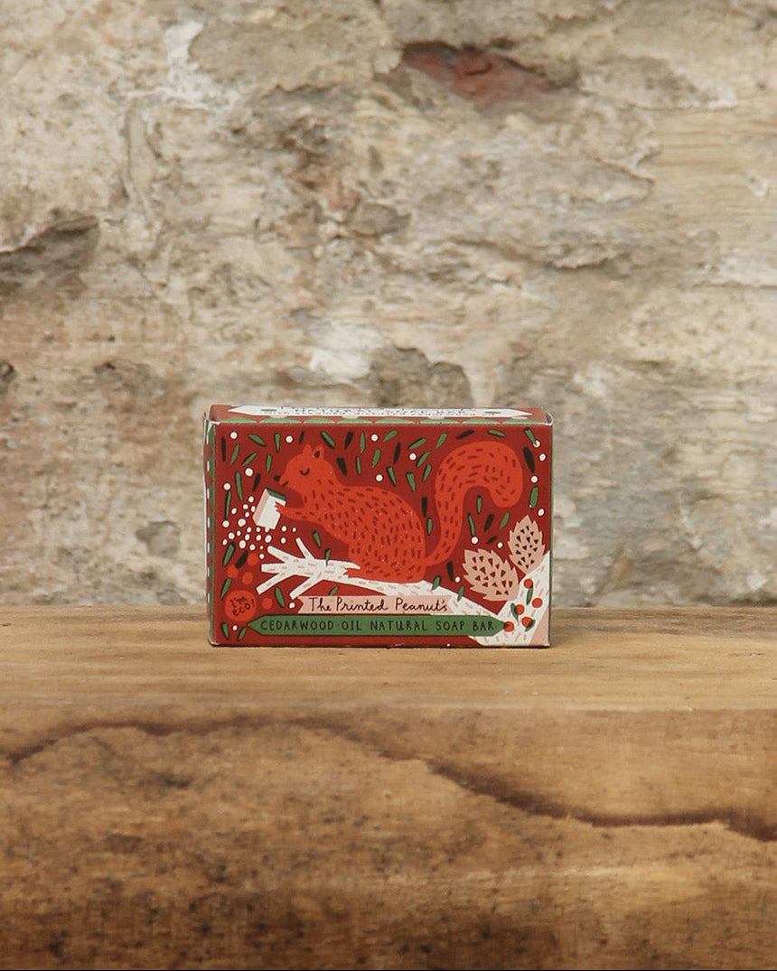 Eine kleine Schachtel von The Printed Peanut | Zedernholz Seife mit einem roten Eichhörnchen und floralen Motiven steht auf einer Holzfläche mit einer strukturierten Steinwand im Hintergrund.