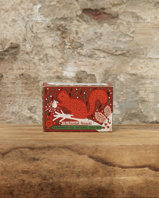 Eine kleine Schachtel von The Printed Peanut | Zedernholz Seife mit einem roten Eichhörnchen und floralen Motiven steht auf einer Holzfläche mit einer strukturierten Steinwand im Hintergrund.