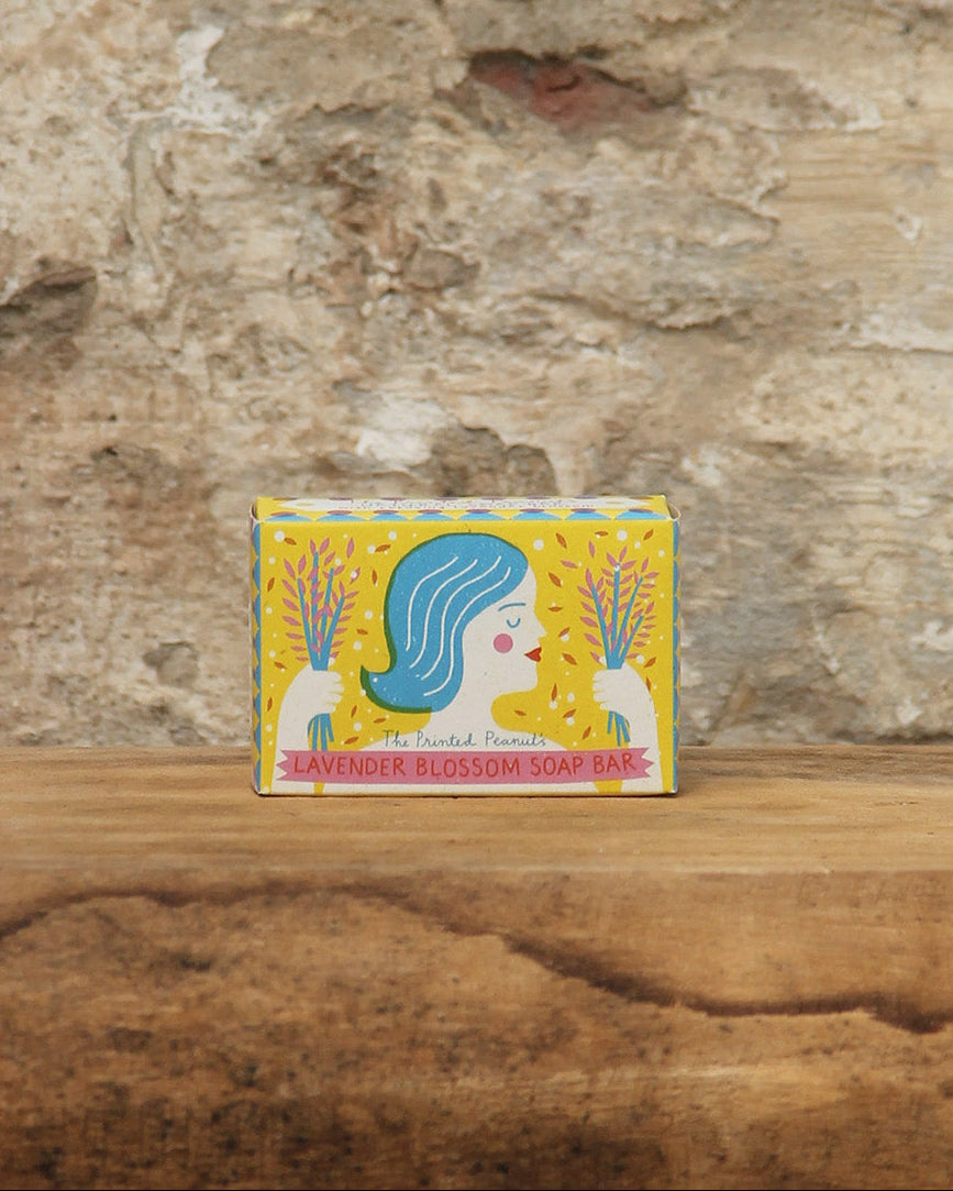 Eine Schachtel der Handseife The Printed Peanut | Lavendelblüte mit einer lebendigen Illustration eines Frauengesichts und Lavendelblüten ruht auf Holz vor einer rustikalen Steinwand.