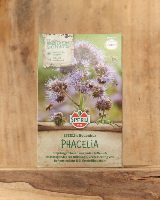 Eine 350g-Packung von Sperli's | Bodenkur 'Phacelia' BIO-Samen zeigt blühende Phacelia-Blüten mit violetten Blütenblättern auf einem Holzhintergrund. Die Verpackung weist auf die Verwendung als Gründünger zur Bodenverbesserung hin.