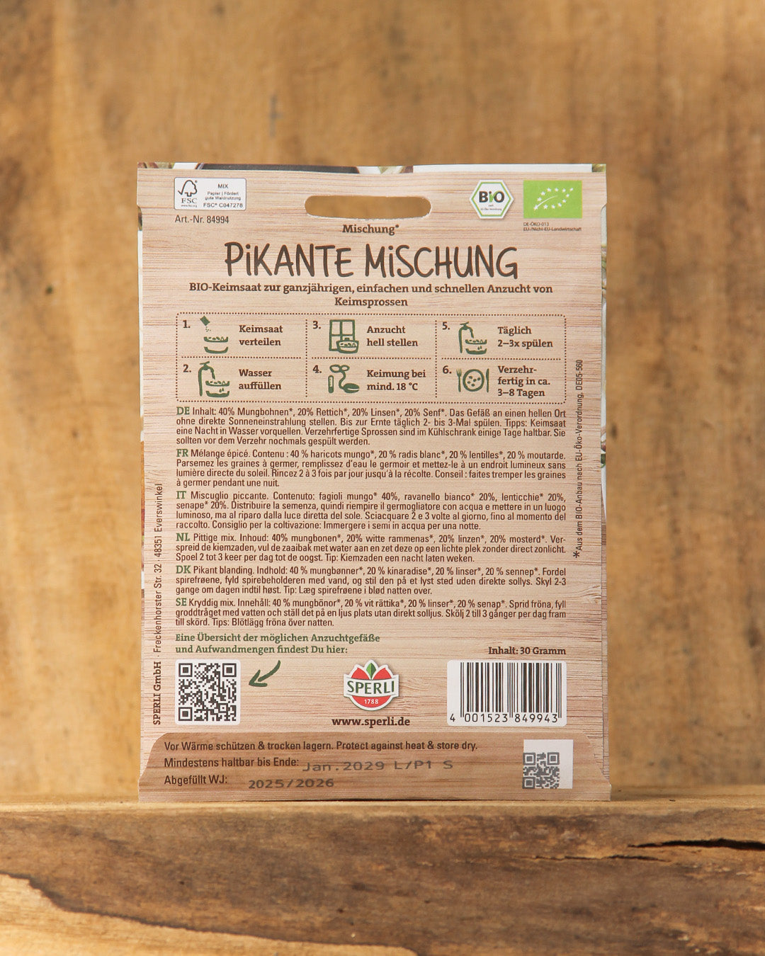 Rückseite einer Sperli's Bio Keimsprossen Saatgut 'Pikante Mischung' Packung mit Pflanzanleitung, deutscher Info mit Symbolen, Bio-Zertifizierungsetiketten und Barcode auf Holzuntergrund.