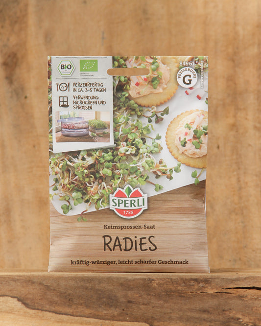 Eine Packung Sperli's Bio Keimsprossen Saat 'Radies' steht aufrecht auf einem Holzbrett mit Bildern von frischen Sprossen, Crackern und deutschem Text und hebt diese bio-zertifizierten Vitaminbomben von Sperli's hervor.