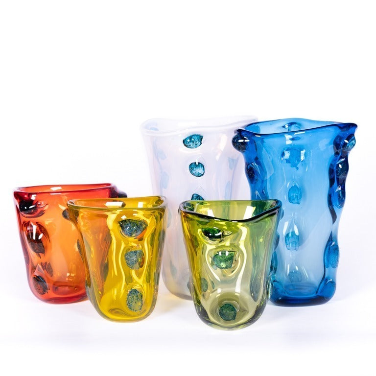Fünf farbenfrohe Glasvasen - rot, gelb, grün, weiß und die Des Pots Skulpturale Vase blau (H 22 cm Ø 20 cm) mit gewellten Rändern und erhabenen Punkten - sind gemeinsam auf weißem Grund angeordnet. Eine auffällige Ergänzung für jede Des Pots-Sammlung.