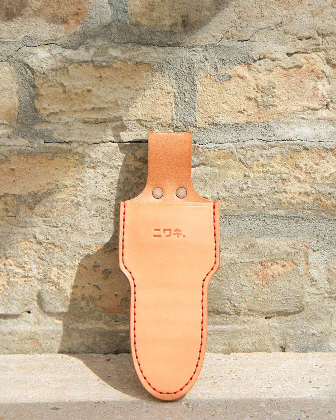 Die Niwaki 'Single Holster' Lederschutzhülle für Schere aus hellbraunem Leder mit roten Nähten und geprägten japanischen Schriftzeichen hängt an einer hellen Backsteinwand, deren Details vom Sonnenlicht hervorgehoben werden.