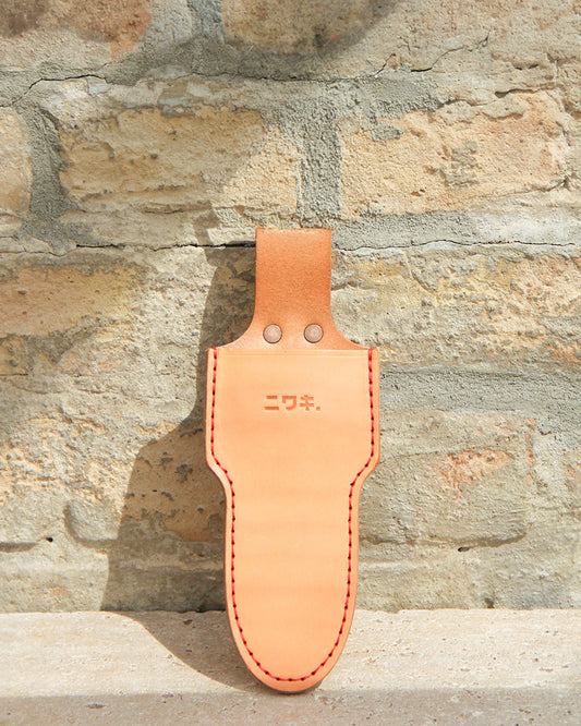 Die Niwaki 'Single Holster' Lederschutzhülle für Schere aus hellbraunem Leder mit roten Nähten und geprägten japanischen Schriftzeichen hängt an einer hellen Backsteinwand, deren Details vom Sonnenlicht hervorgehoben werden.