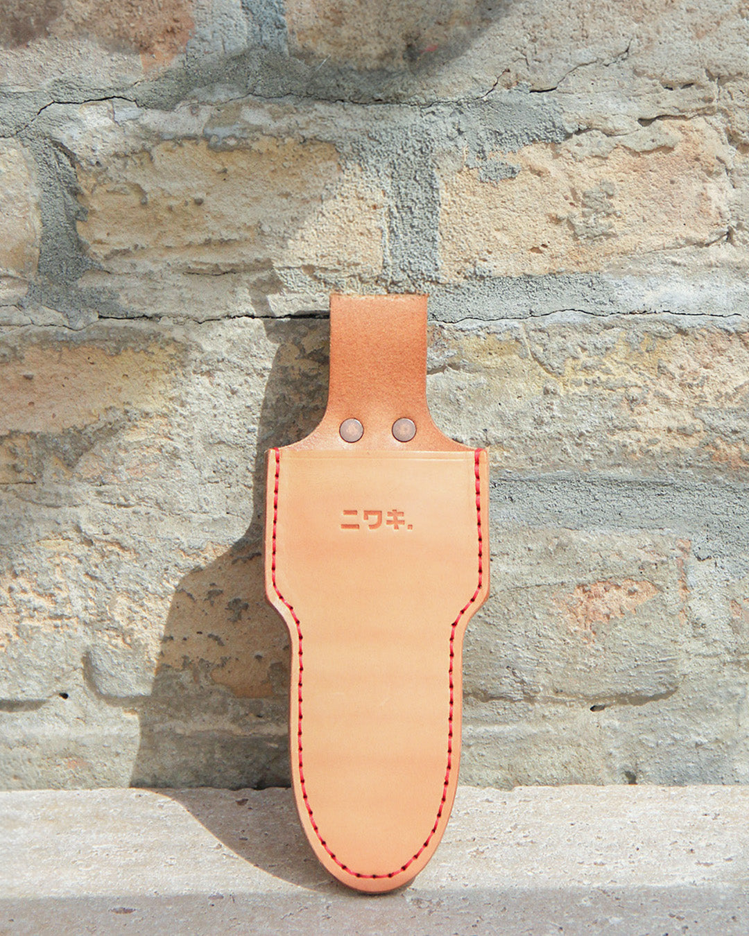 A Niwaki | „Single Holster“ – eine Lederschutzhülle für eine Schere aus hellbraunem Leder, verziert mit roten Nähten und japanischen Schriftzeichen auf der Vorderseite, hängt elegant an einer rauen, hellen Backsteinwand.