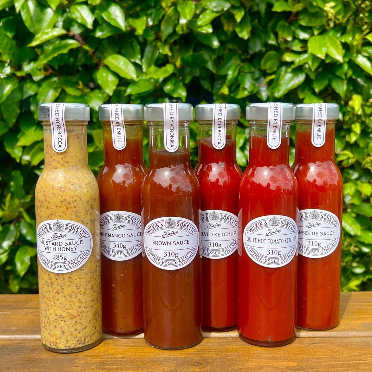 Sechs Glasflaschen mit verschiedenen Saucen von Wilkin & Sons, darunter die Tiptree Honig-Senf-Sauce (260 ml), stehen in einer Reihe auf einer Holzunterlage. Jede Flasche hat einen silbernen Verschluss und ein weißes Etikett und enthält Sorten wie Ketchup und Senf. Dahinter bilden üppige, grüne Blattpflanzen einen lebendigen Hintergrund.
