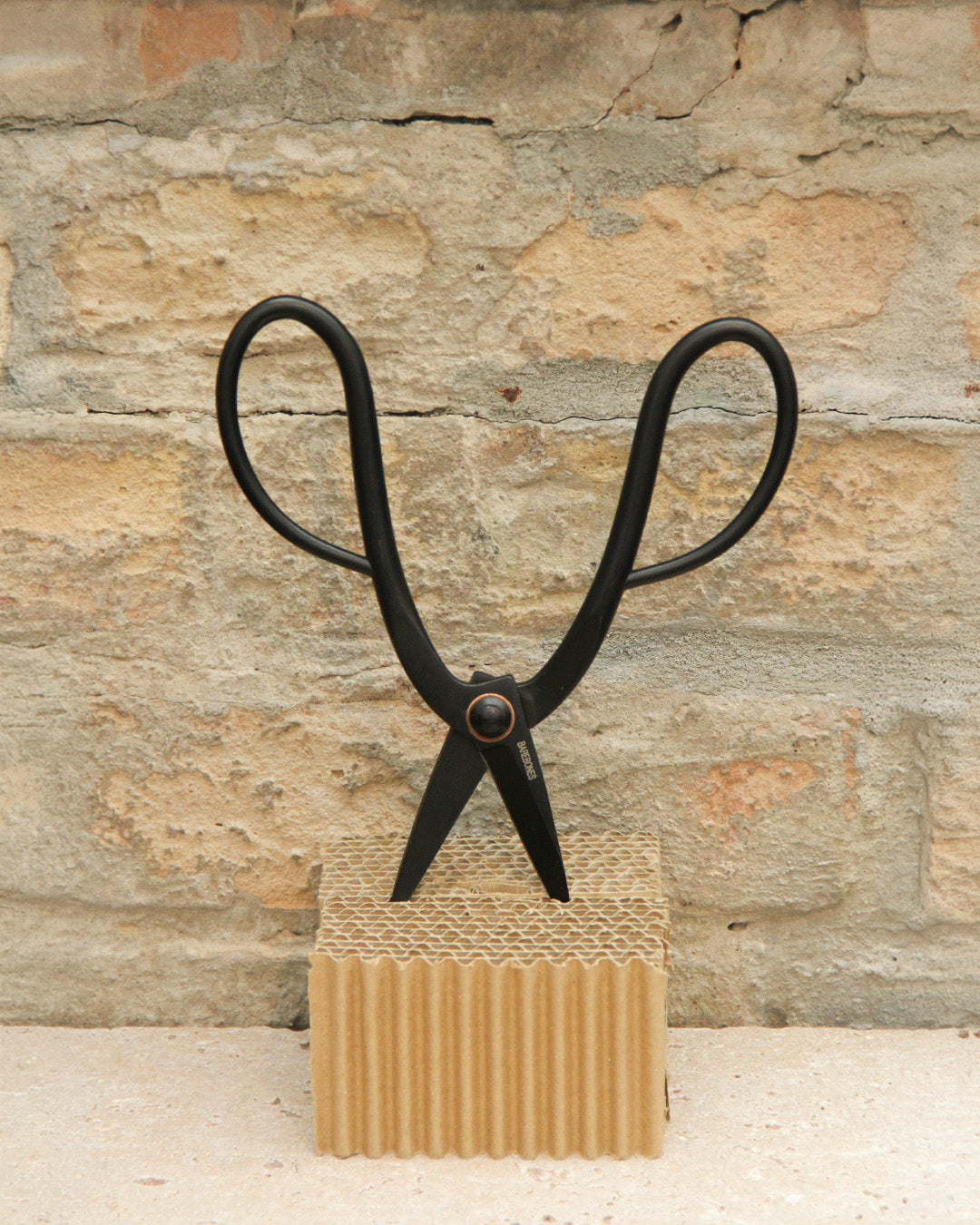 Ein Paar Barebones | Artisan Gartenschere aus Edelstahl mit großen Schlaufengriffen steht aufrecht auf Wellpappe vor dem Hintergrund einer strukturierten Steinwand.