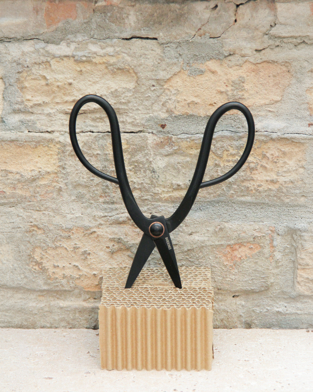 Die „Barebones“ | Eine handgefertigte Gartenschere aus Edelstahl mit großen Scherenbögen und einer eleganten schwarzen Oberfläche steht aufrecht auf einem Stück Wellpappe vor einer strukturierten Steinwand.