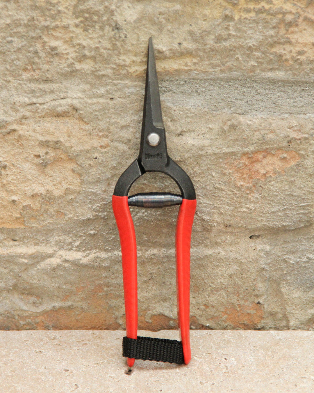Die Blumenschere Niwaki | 'Forged Snips' mit roten Griffen und spitzen Klingen steht aufrecht an einer strukturierten Steinwand. Ein schwarzes Band in der Nähe der Griffe zeigt die präzise Handwerkskunst dieser japanischen Gartenschere von Niwaki.