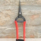 Ein Paar Niwaki | „Forged Snips“-Blumenscheren von Niwaki mit roten Griffen und einer schwarzen Metallklinge, aufrecht vor dem Hintergrund einer Steinmauer platziert.