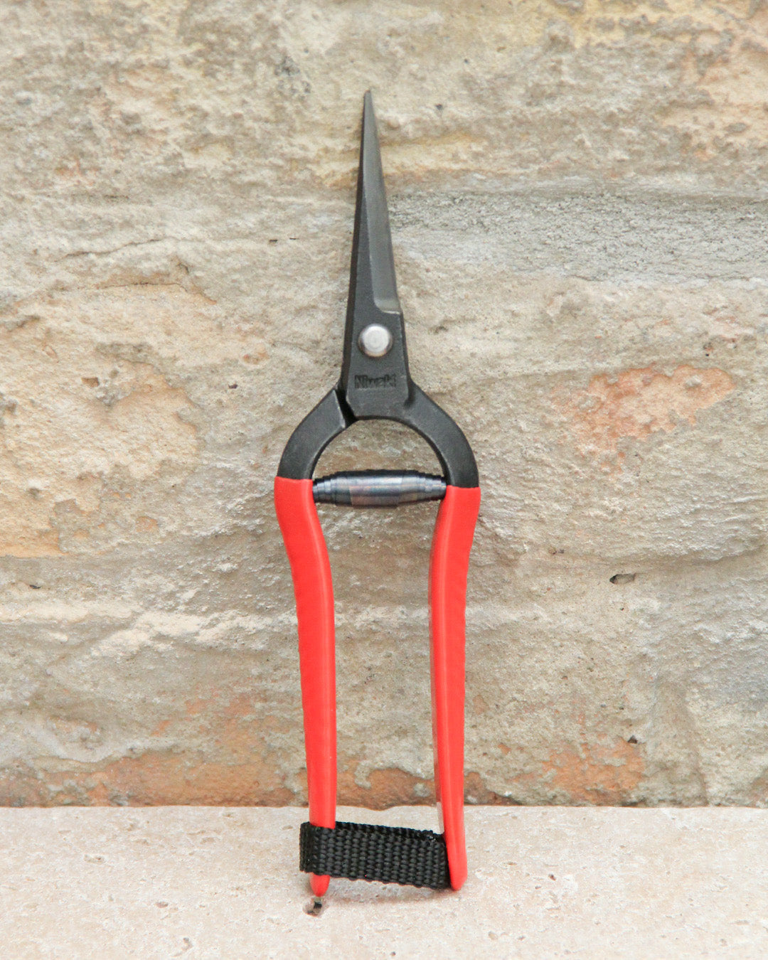 Ein Paar Niwaki | „Forged Snips“-Blumenscheren von Niwaki mit roten Griffen und einer schwarzen Metallklinge, aufrecht vor dem Hintergrund einer Steinmauer platziert.