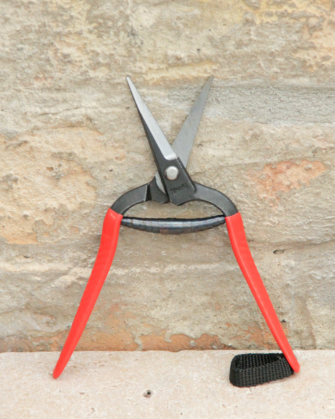 Ein Paar „Forged Snips“-Blumenscheren von Niwaki mit roten Griffen, einem schwarzen Griffbereich und einem schwarzen Riemen an einem der Griffe, lehnt mit geöffneten Klingen an einer strukturierten Steinmauer.