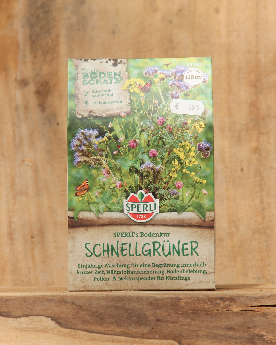 Eine Packung Sperli's | Bodenkur 'Schnellgrüner' 450 g steht aufrecht auf einer Holzunterlage mit bunten Wildblumen-Illustrationen und deutschem Text über diesen schnell wirkenden Bodenverbesserer von Sperli's.