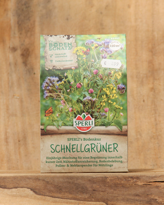 Eine Packung Sperli's | Bodenkur 'Schnellgrüner' 450 g steht aufrecht auf einer Holzunterlage mit bunten Wildblumen-Illustrationen und deutschem Text über diesen schnell wirkenden Bodenverbesserer von Sperli's.