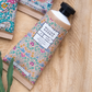 Eine Tube William Morris at Home | 'Eyebright' Handcreme 50 ml mit einem floral gemusterten Etikett ruht auf einer Holzfläche neben einem grünen Blatt und anderen dekorativen Tuben.