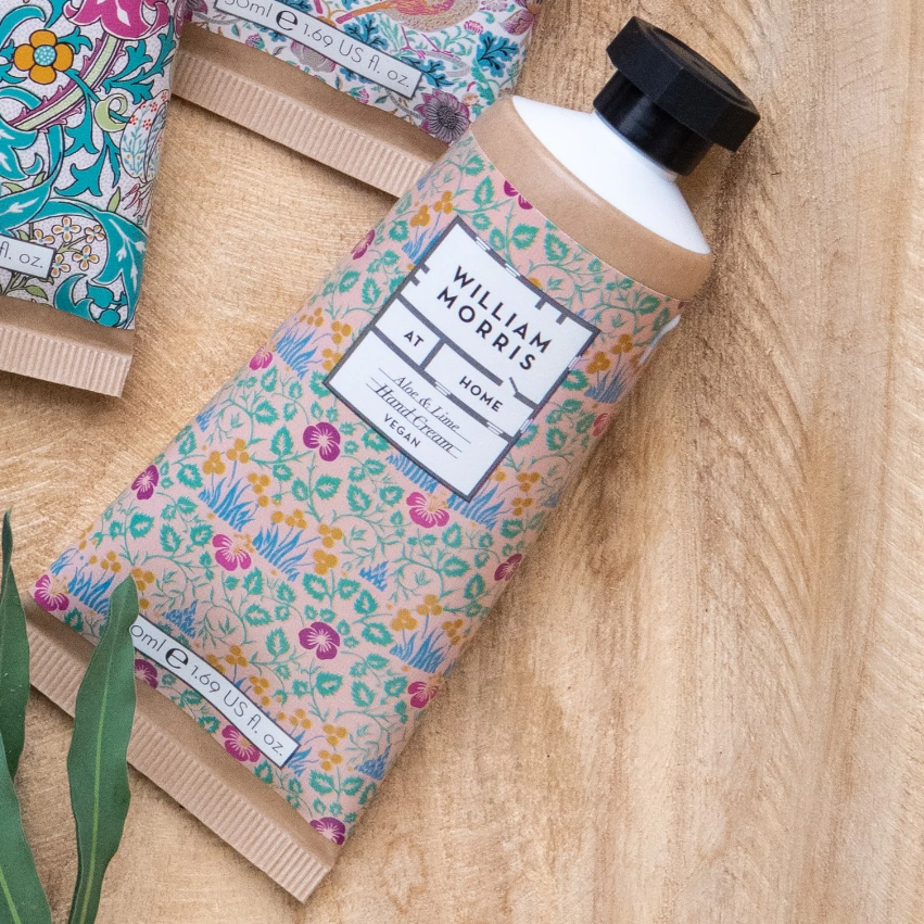 Eine Tube William Morris at Home | 'Eyebright' Handcreme 50 ml mit einem floral gemusterten Etikett ruht auf einer Holzfläche neben einem grünen Blatt und anderen dekorativen Tuben.