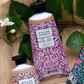 Eine Tube William Morris at Home 'Acorn' Handcreme 50 ml mit einem lila Blumenetikett ruht zwischen grünen Blättern und Blumen auf beigem Stoff.