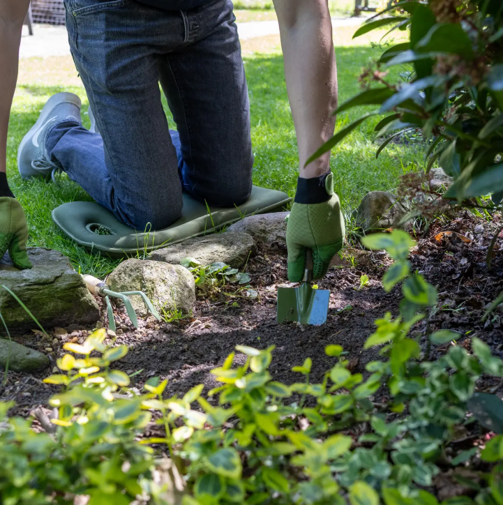 Eine Person mit grünen Handschuhen kniet auf dem Esschert Kniekissen | Memory-Schaum grün und benutzt eine kleine Handschaufel, um den Garten zu pflegen, was die Gartenarbeiten an einem sonnigen Tag inmitten von Pflanzen und Steinen bequemer und einfacher macht.