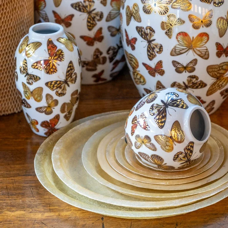 Die Des Pots Porzellan Vase mit Schmetterlingen (H 11 Ø 11) von Des Pots steht auf einem Holztisch neben goldfarbenen, runden Tellern.