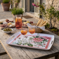 Im Außenbereich ist ein rustikaler Holztisch mit dem Tablett "Cecile" (46 x 36 cm) von Napking gedeckt, das mit einem floralen Leinendruck verziert ist und auf dem orangefarbene Cocktails, ein Getränkekrug, Brotstangen, Oliven und Wurstwaren liegen. Steinmauern und Blumen vervollständigen die Szene.