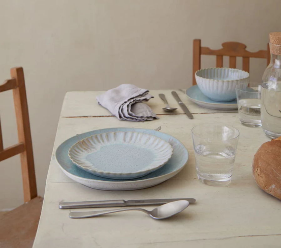 Ein rustikaler Tisch ist mit Costa-Nova-Salattellern in "Mallorca-Meeresblau", einem Glas Wasser, Besteck, einer Schüssel, Leinenservietten, einem Brotlaib und einer Glaswasserflasche mit Korkdeckel gedeckt. Zwei Holzstühle sind ebenfalls zu sehen.