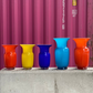 Fünf leuchtende Vasen von Des Pots sind auf Beton vor einem magentafarbenen Schiffscontainer aufgereiht. Der Star ist die Des Pots | Blaue Vase aus Glas in Kelchform H 18 cm Ø 11 cm, die zwischen orangen, gelben und türkisfarbenen Gefährten hervorsticht.