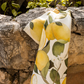 Das Napking Premium Leinen Geschirrtuch 'Amalfi' (55 × 75 cm) zeigt einen lebendigen Zitronen- und Blätterdruck, der über eine rustikale Steinmauer im Sonnenlicht drapiert ist, mit üppigem Grün im Hintergrund.