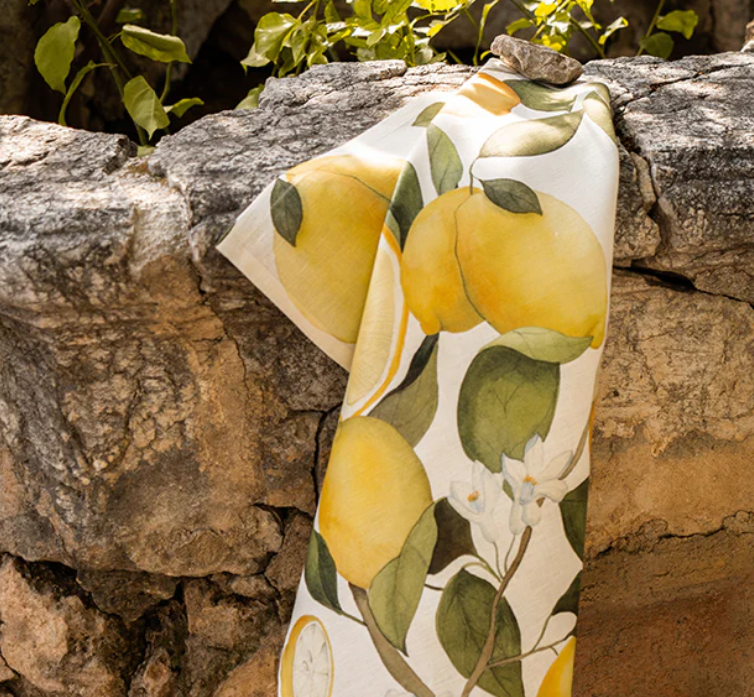 Das Napking Premium Leinen Geschirrtuch 'Amalfi' (55 × 75 cm) zeigt einen lebendigen Zitronen- und Blätterdruck, der über eine rustikale Steinmauer im Sonnenlicht drapiert ist, mit üppigem Grün im Hintergrund.