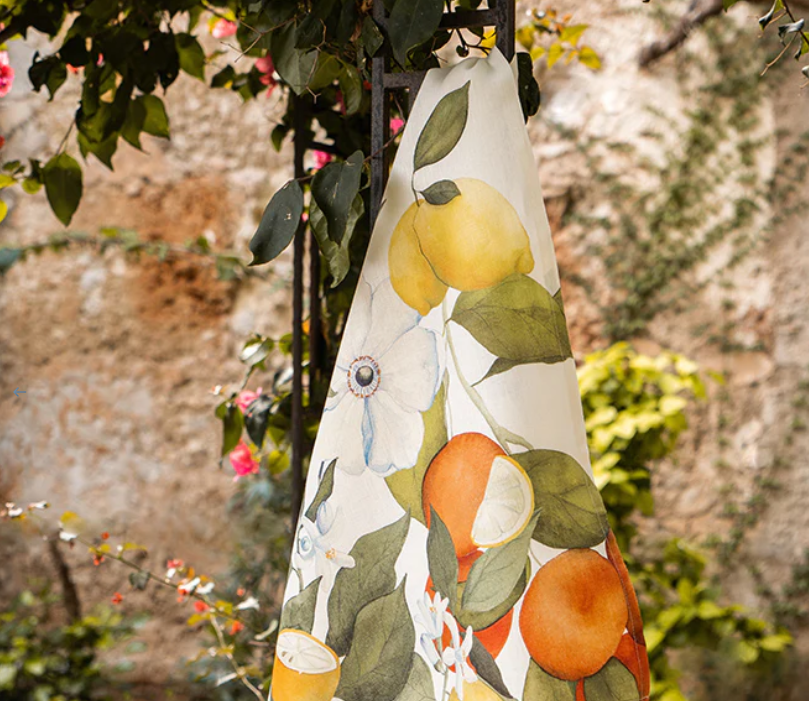 Das Napking | Premium Leinen Geschirrtuch 'Amalfi' (55 × 75 cm) zeigt ein lebhaftes Zitrusfrucht- und Blumendesign und bringt mediterranen Charme in Ihre Küche. Das Geschirrtuch hängt im Freien an einer rustikalen Steinwand. Marke: Napking.