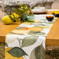 Ein Holztisch für draußen mit dem Napking Premium Leinen Tischläufer 'Amalfi' 50 x 150 cm, grünen Gläsern, Schalen, Tellern und frischen Zitronen mit grünen Blättern.