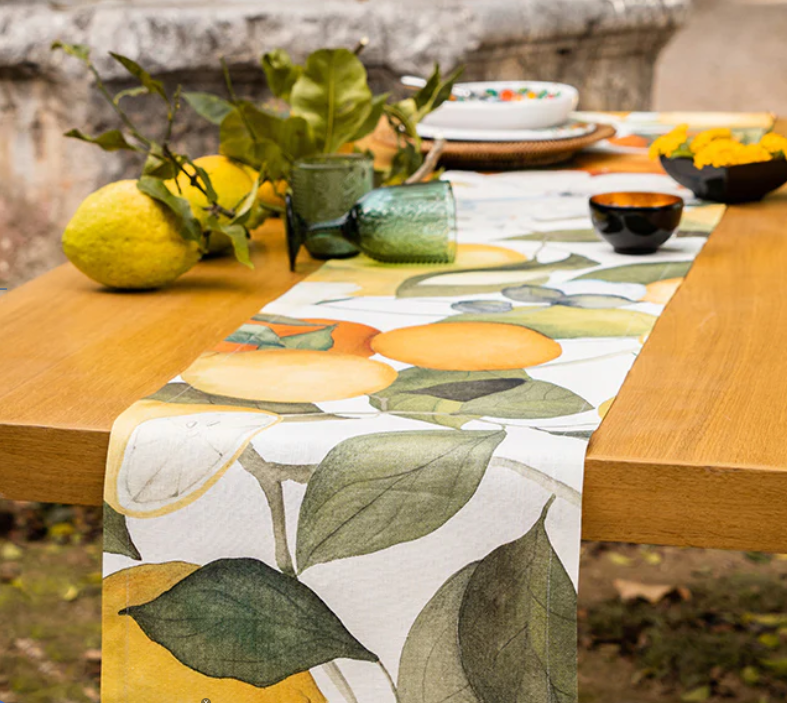 Ein Holztisch für draußen mit dem Napking Premium Leinen Tischläufer 'Amalfi' 50 x 150 cm, grünen Gläsern, Schalen, Tellern und frischen Zitronen mit grünen Blättern.