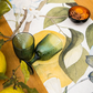 Zwei grüne Gläser stehen auf dem Napking | Premium Leinen Tischläufer 'Amalfi' 50 x 150 cm, der ein frisches botanisches Design aufweist. Eine schwarze Schale mit getrockneten Aprikosen und Mandeln steht daneben, während Zitronenzweige den Holztisch schmücken.
