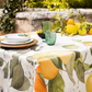 Auf einem Tisch im Freien steht die Napking Premium Leinen Tischdecke 'Amalfi' 110 x 110 cm von Napking, gepaart mit grünen Gläsern, einer weißen Blumenschale, Silberbesteck, Zitrusfrüchten und Grünzeug, vor einer Steinwand und Pflanzen.