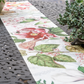 Die Nahaufnahme eines Tisches im Freien zeigt den Napking Premium Leinen Tischläufer 'Dafne' 50 x 150 cm und eine eingetopfte Sukkulente auf der rechten Seite, die das elegante florale Muster des Läufers von Napking hervorhebt.