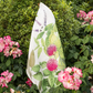 Das Napking | Premium Leinen Geschirrtuch 'Épices' (55 × 75 cm), verziert mit farbenfrohen botanischen Drucken, drapiert sich elegant über einen Busch inmitten von rosa und weißen Blüten und bringt mediterrane Eleganz in den Garten.