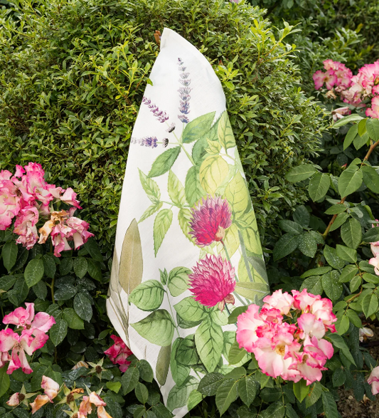 Das Napking | Premium Leinen Geschirrtuch 'Épices' (55 × 75 cm), verziert mit farbenfrohen botanischen Drucken, drapiert sich elegant über einen Busch inmitten von rosa und weißen Blüten und bringt mediterrane Eleganz in den Garten.