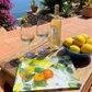 Auf einer sonnigen Terrasse mit Meerblick und mediterranen Pflanzen steht ein Napking Tablett 'Amalfi' 46 x 36 cm mit einer Flasche Weißwein, zwei gefüllten Gläsern und einer blauen Schale mit Zitronen auf einem Holztisch.