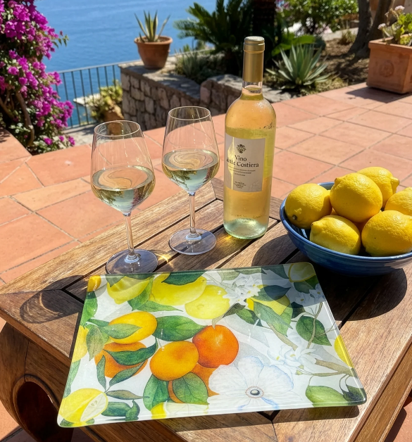 Auf einer sonnigen Terrasse mit Meerblick und mediterranen Pflanzen steht ein Napking Tablett 'Amalfi' 46 x 36 cm mit einer Flasche Weißwein, zwei gefüllten Gläsern und einer blauen Schale mit Zitronen auf einem Holztisch.