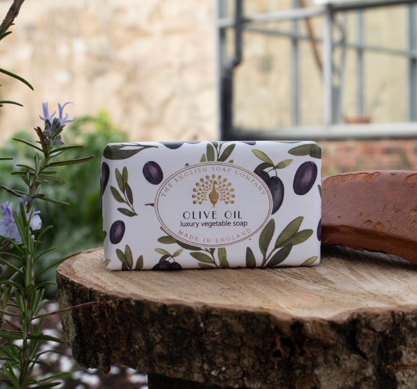 Eine ESC | 'Olive' Luxuriöse Olivenölseife mit mediterranem Duft von The English Soap Company liegt in olivenverziertem Papier verpackt auf einem Baumstumpf im Freien neben einer Pflanze und einem Ziegelstein.