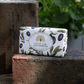 Eine Tafel ESC | 'Olive' Luxuriöse Olivenölseife mit mediterranem Duft von The English Soap Company, in Papier mit Olivenmotiven gewickelt, liegt draußen auf einem Ziegelstein vor grünem Hintergrund.