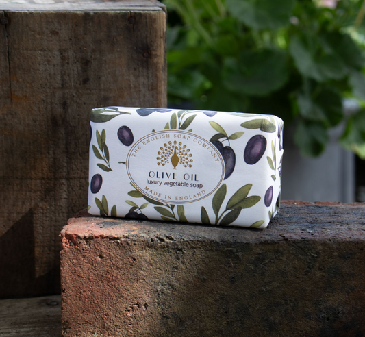 Eine Tafel ESC | 'Olive' Luxuriöse Olivenölseife mit mediterranem Duft von The English Soap Company, in Papier mit Olivenmotiven gewickelt, liegt draußen auf einem Ziegelstein vor grünem Hintergrund.