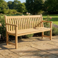 Die Alexander Rose | 'Roble Wood Broadfield Bench' Gartenbank 122 cm aus edlem Roble-Holz steht auf einer Steinterrasse, umgeben von blühenden Blumen, Bäumen und Rasen im Sonnenlicht.