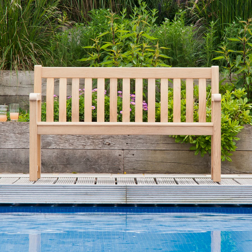 Die wetterfeste Gartenbank „Roble St Georges Bench“ von Alexander Rose (150 cm) steht auf einer Terrasse am blauen Pool, im Hintergrund sind grüne Pflanzen und eine Stützmauer aus Holz zu sehen. Am Rand steht ein Glas mit einem orangefarbenen Getränk.